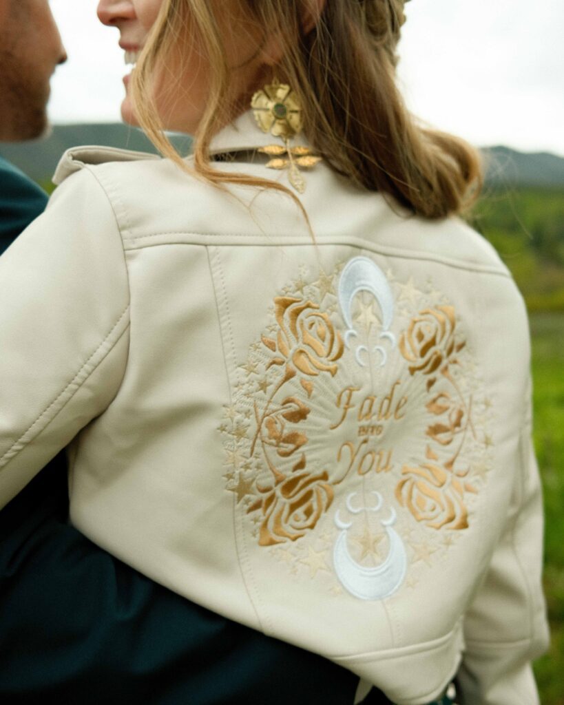 Erin wearing her custom embroidered vegan leather jacket that says "fade into you" in gold with gold and white floral and celestial details while embracing josh