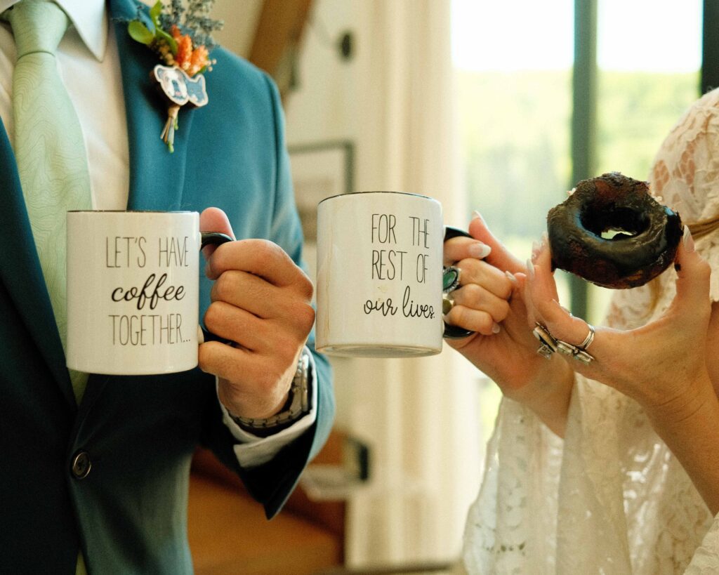 erin and josh drinking coffee and eating vegan chocolate covered donuts with mugs that say "lets have coffee together for the rest of our lives"